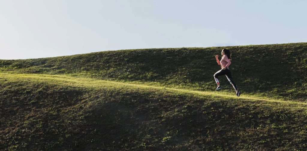 Frau läuft einen grasbewachsenen Hügel hinauf, Symbol für HIIT-Training im Beitrag zu Hashimoto und Sport.